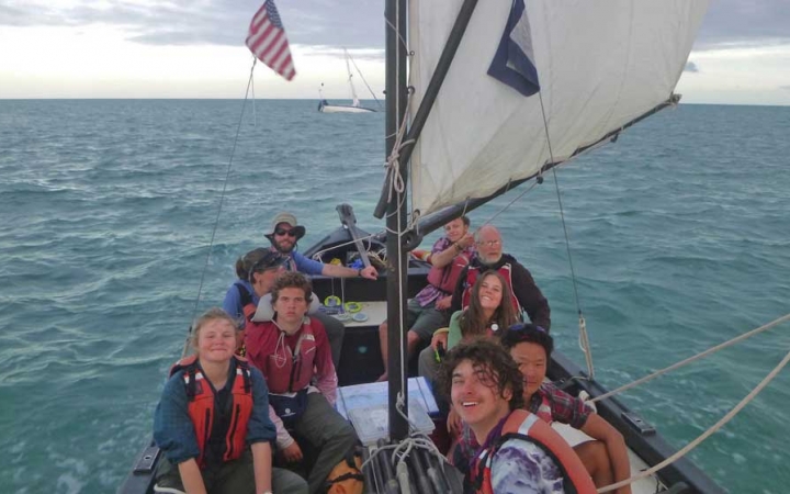 A group of young people sitting in a sailboat on calm. blue water smile at the camera.